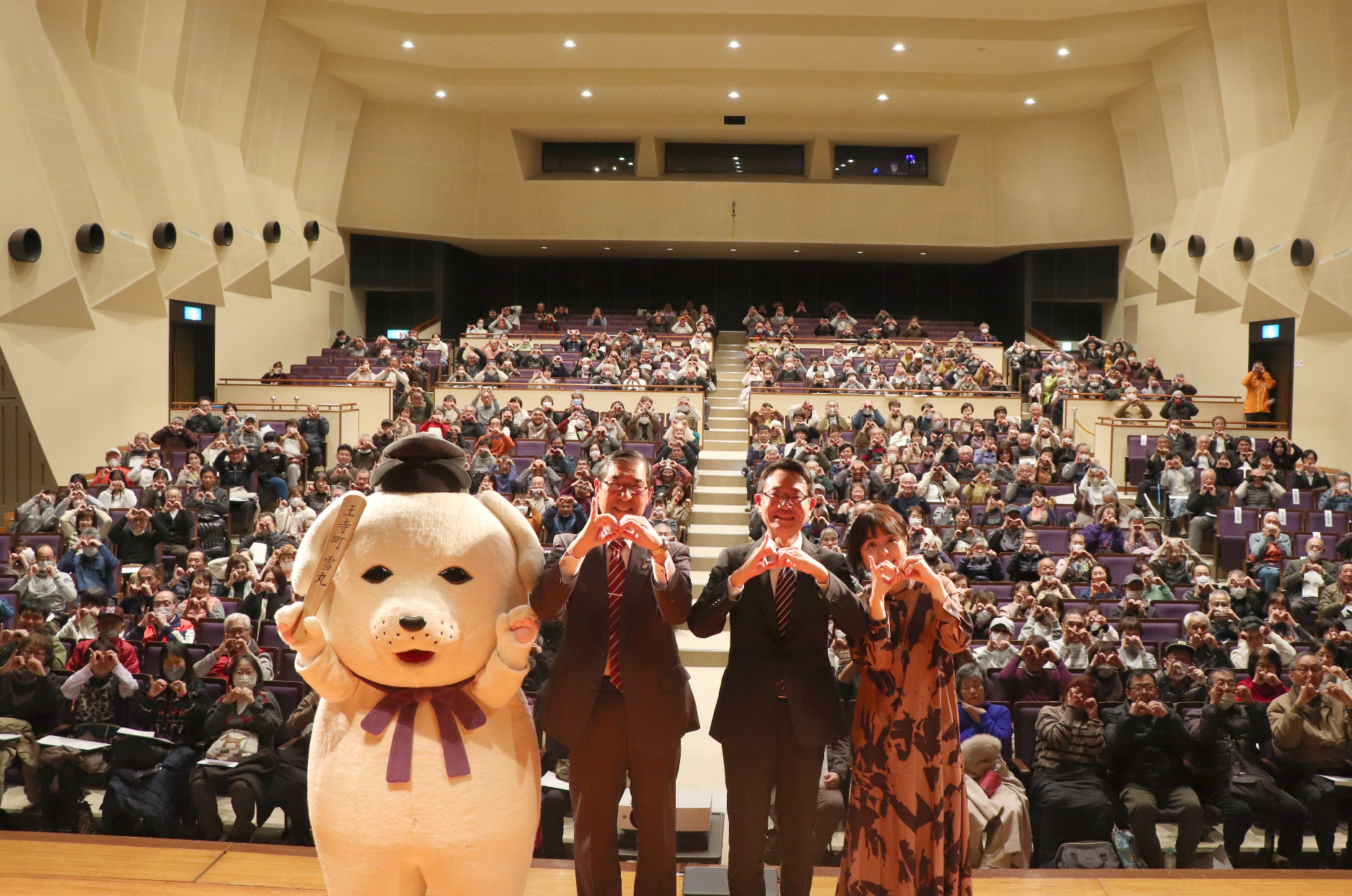 客席をバックにひゃくまるポーズをする登壇者3人（平井町長・野村明大氏、中元綾子氏、雪丸）